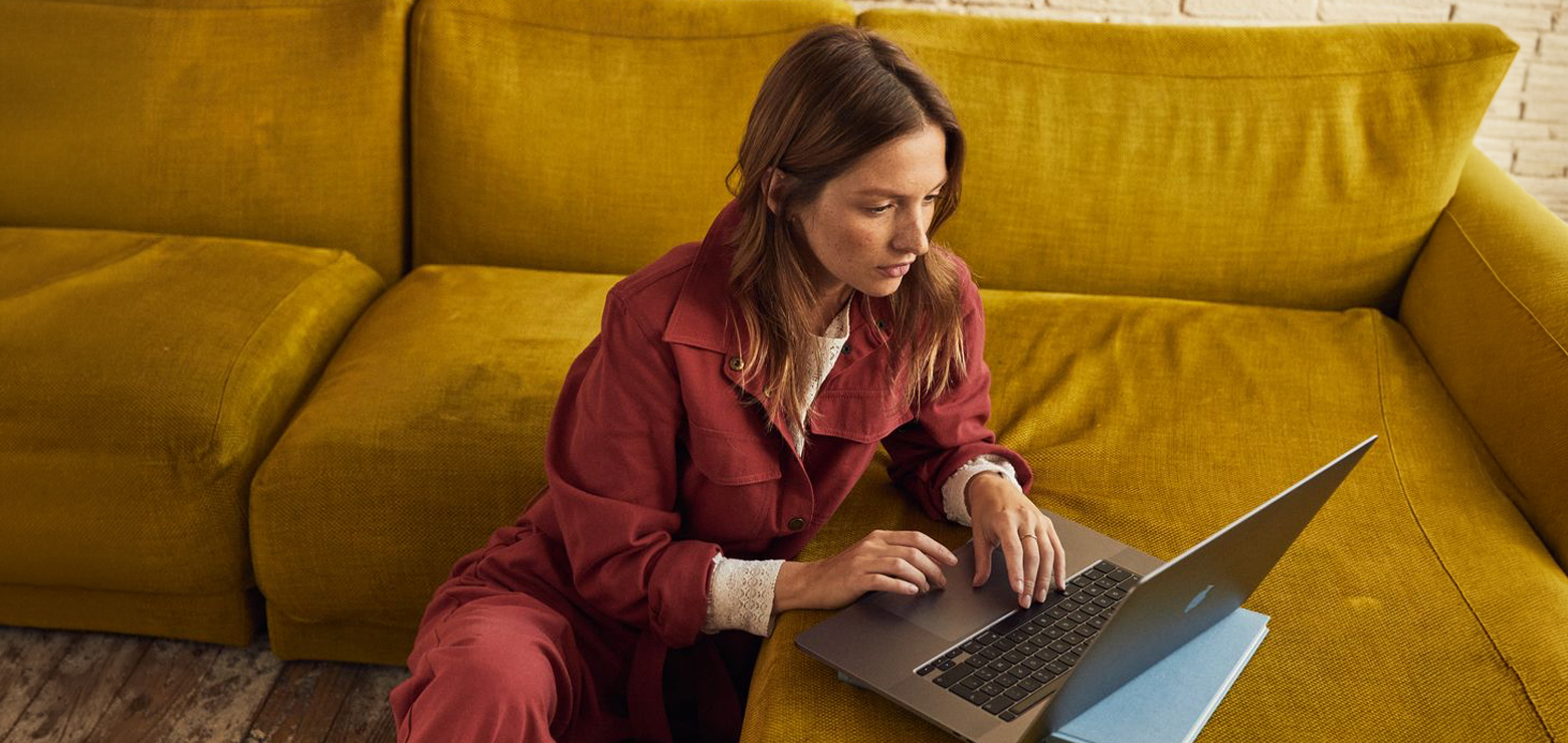 A woman in a rust-red jumpsuit sitting on the floor and leaning against a mustard-yellow couch while focused on her laptop.