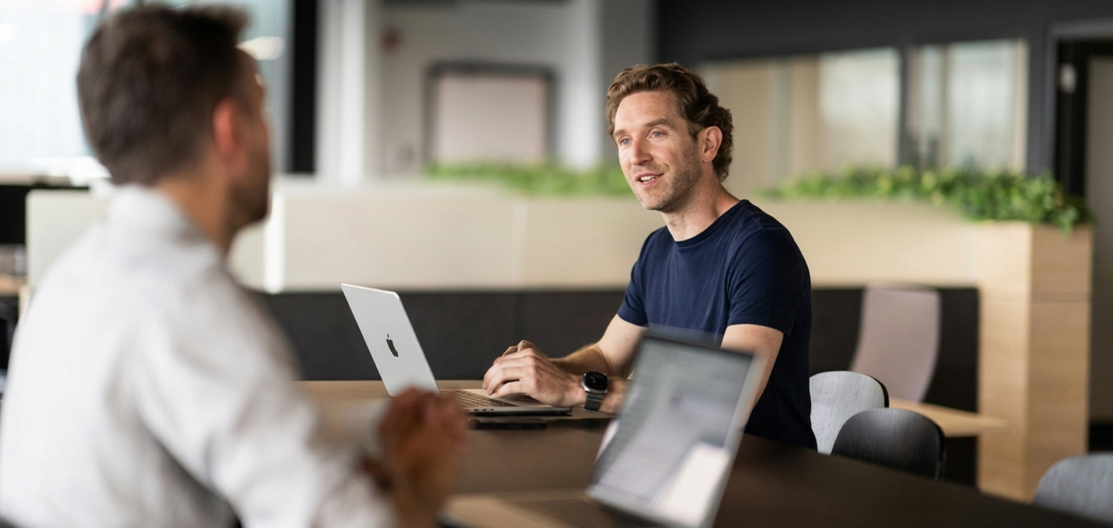 Two businessmen in a modern corporate office having a professional discovery discussion across a dark conference table with open laptops, representing an evaluation framework for hiring an AI visibility agency in the USA