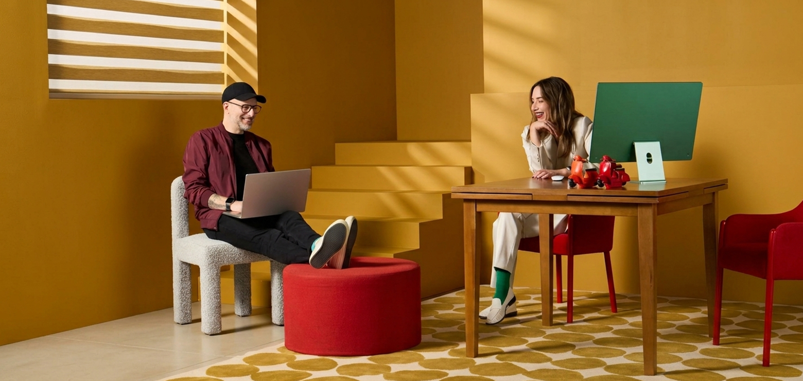 A modern, stylized tech office with vibrant yellow walls, featuring a man with a laptop sitting on a white bouclé chair with feet resting on a red ottoman, collaborating with a woman at a wooden desk with a green computer monitor