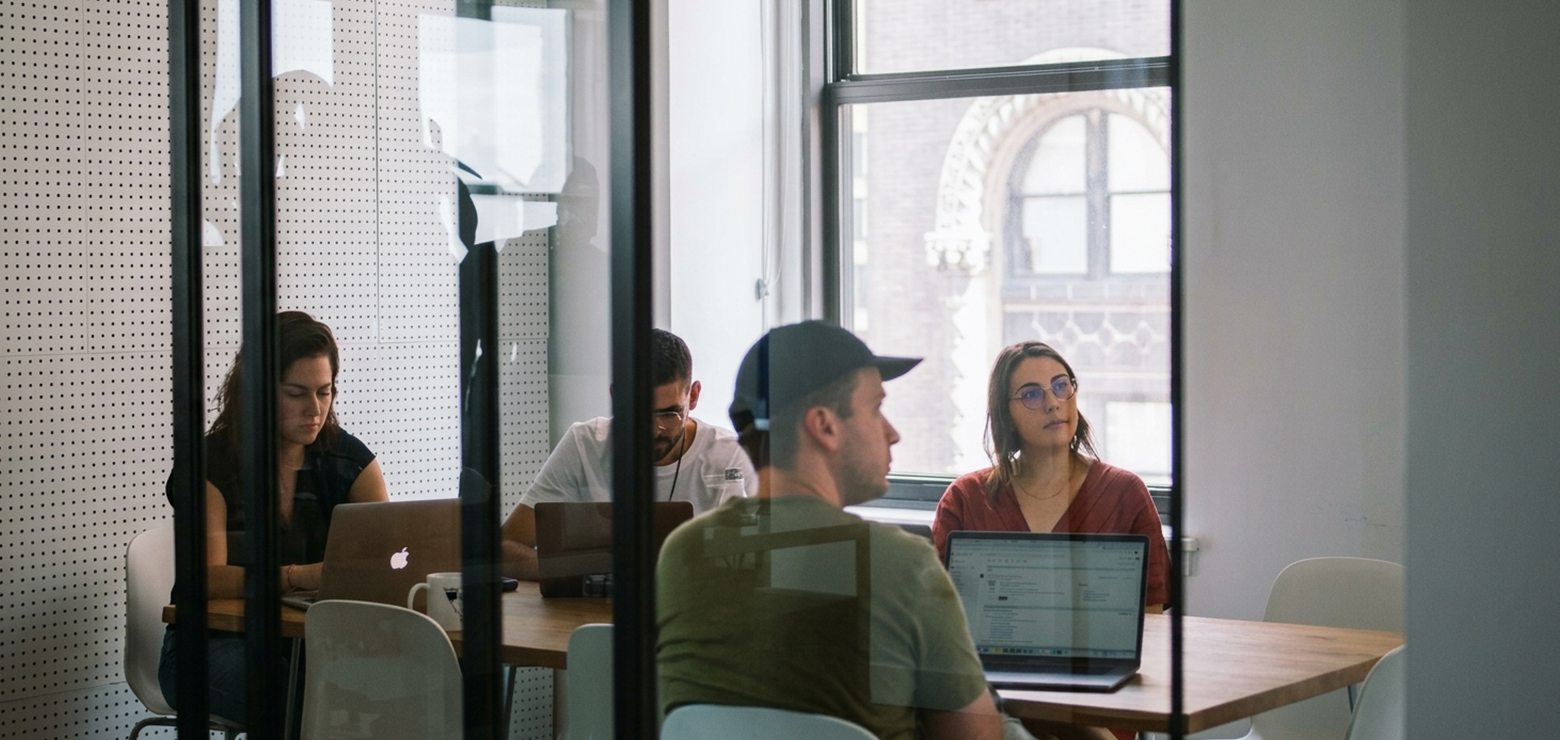 A corporate marketing team working on laptops around a wooden conference table, seen through a glass office partition, illustrating an enterprise transition from single-channel SEO to AI growth engines
