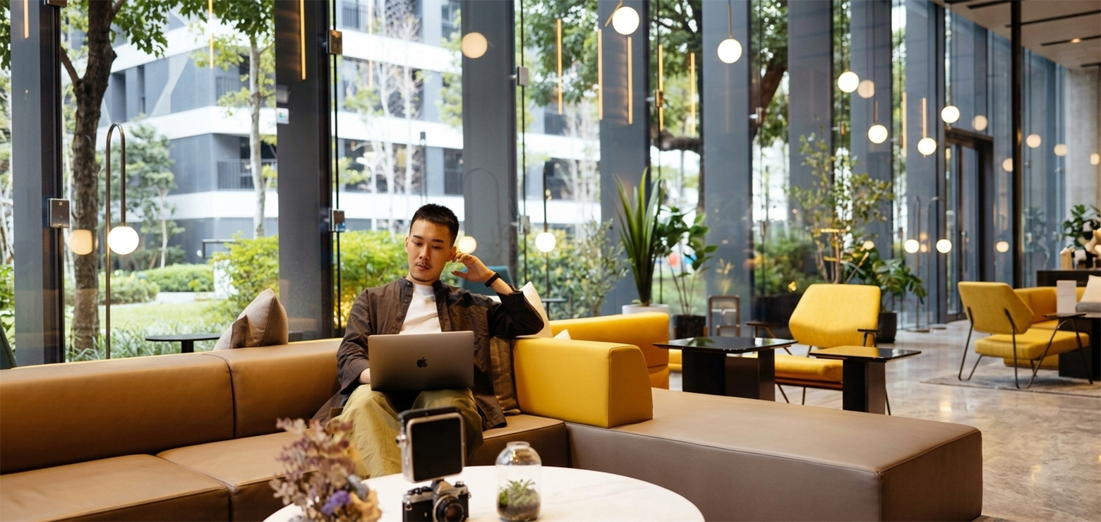 A professional man sitting on a modern brown and yellow modular sofa in a spacious, sunlit glass lobby while working on a laptop, representing content strategy creation in a modern corporate workspace.