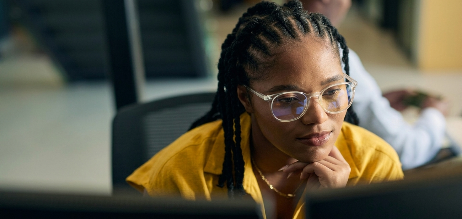 A close-up of a data analyst with braided hair wearing clear-rimmed glasses and a yellow top, leaning forward and focusing deeply on dual computer monitors in a corporate office setting.