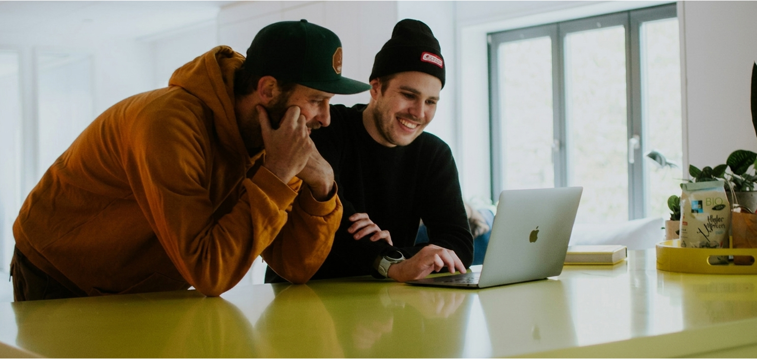 Two smiling male colleagues in casual tech attire, one wearing a hoodie and cap, the other a beanie, leaning over a laptop placed on a bright lime-green counter to review AI search rankings and data analytics