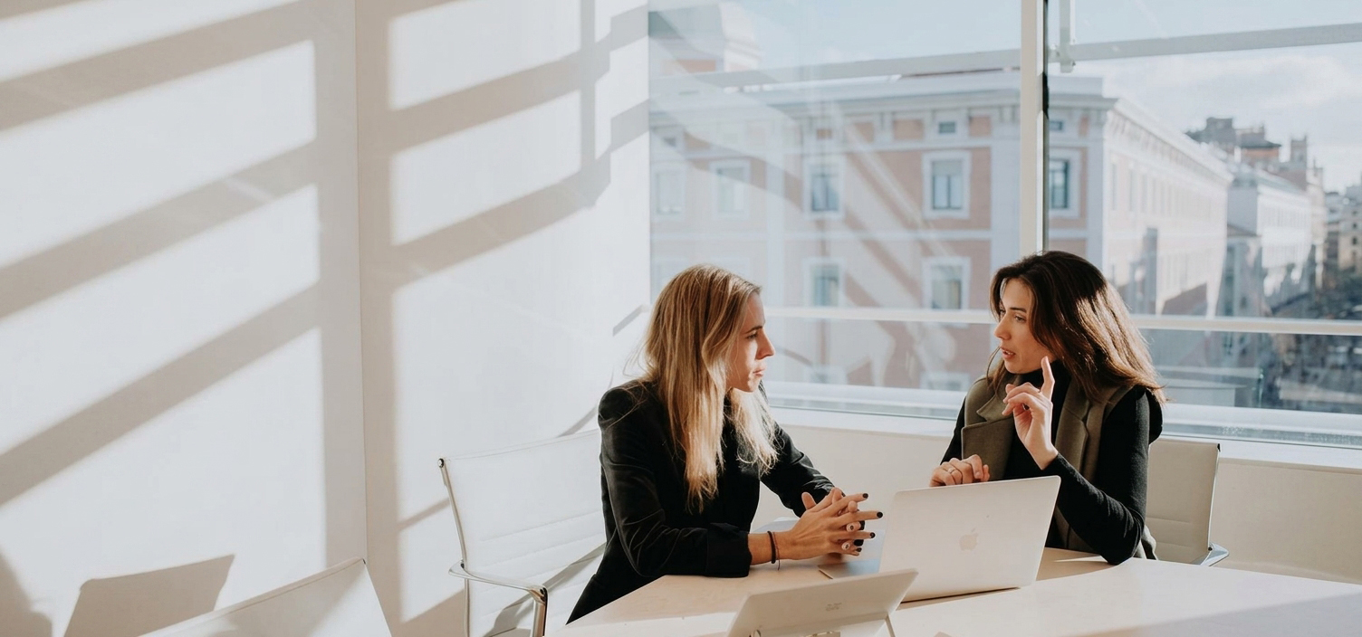 Two businesswomen sitting at a conference table with open laptops in a bright, sunlit modern office, engaged in a serious discussion about AI data security policies and protecting brand content from unauthorized machine learning training.