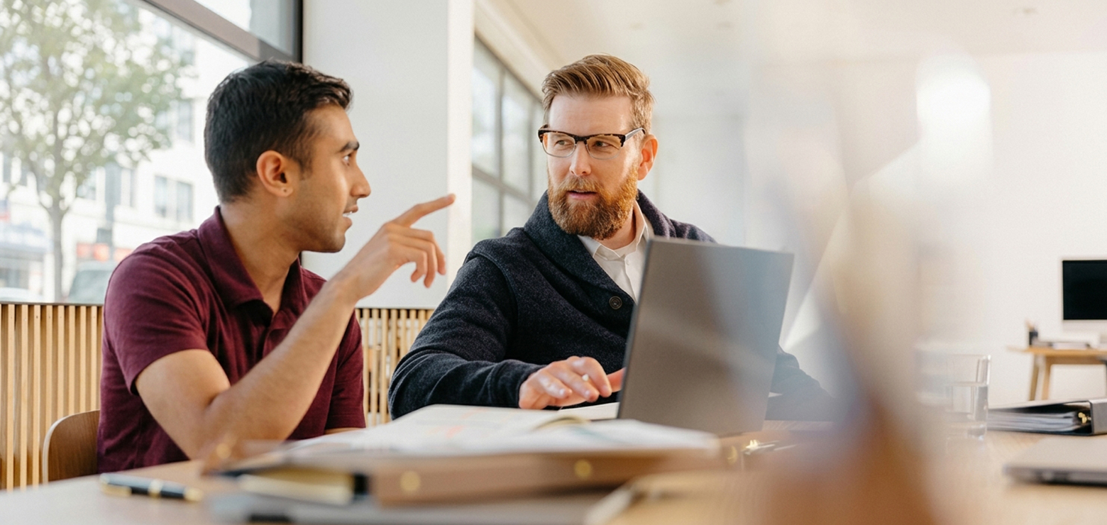 A modern strategy meeting between two male professionals in a sunlit office, with one pointing at a laptop screen while reviewing AI visibility attribution metrics and share-of-voice data for brand mentions