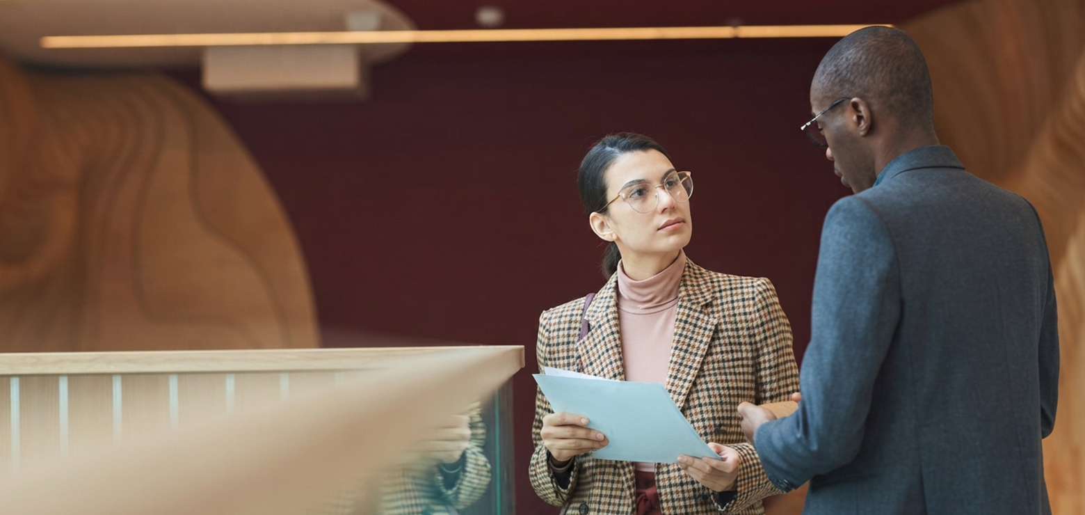 A female corporate executive in a plaid blazer and glasses holding audit reports while discussing Google Tag updates and revenue attribution leaks with a male colleague in a modern office atrium