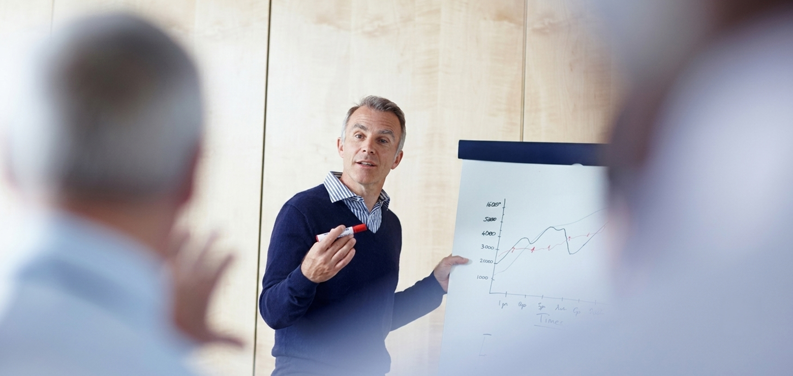 A senior male speaker in a blue sweater presenting growth charts on a flipchart to a group of executives, demonstrating underrated AI SEO strategies that drive organic traffic