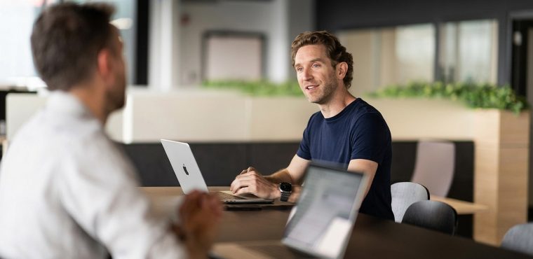 Two businessmen in a modern corporate office having a professional discovery discussion across a dark conference table with open laptops, representing an evaluation framework for hiring an AI visibility agency in the USA