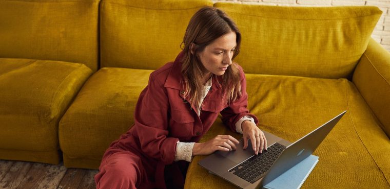 A woman in a rust-red jumpsuit sitting on the floor and leaning against a mustard-yellow couch while focused on her laptop.