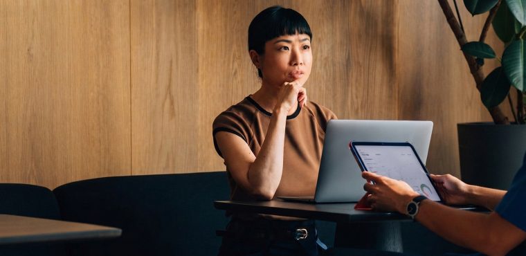 An Asian female executive in a brown top sitting thoughtfully at a laptop while reviewing analytics data displayed on a tablet during a business consultation, illustrating agentic commerce discoverability and GEO strategies for autonomous AI search