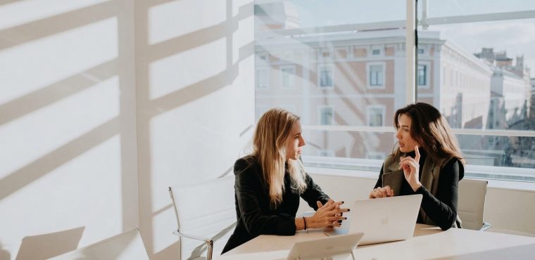 Two businesswomen sitting at a conference table with open laptops in a bright, sunlit modern office, engaged in a serious discussion about AI data security policies and protecting brand content from unauthorized machine learning training.