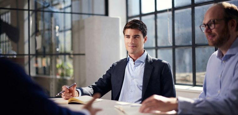 A professional business meeting featuring a consultant in a dark suit jacket holding a pen while listening attentively to a client across a conference table, illustrating AI search optimization strategies for local businesses