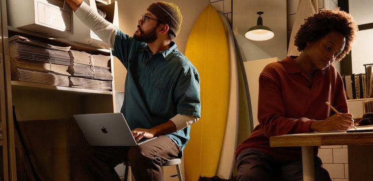 A male creative strategist in a beanie using a MacBook Air while reaching for file boxes on storage shelves alongside a Black female colleague taking notes at a wooden table in a Southern California studio with surfboards, illustrating a benchmark of California GEO agencies