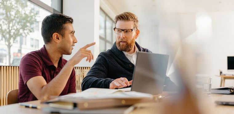 A modern strategy meeting between two male professionals in a sunlit office, with one pointing at a laptop screen while reviewing AI visibility attribution metrics and share-of-voice data for brand mentions