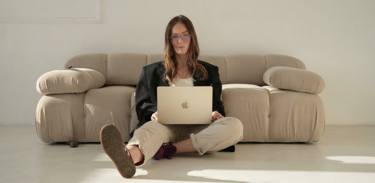 A female enterprise marketing strategist with glasses wearing a black blazer and beige pants sitting on the floor leaning against a beige couch working on a gold laptop, illustrating entity citations tracking in generative engines