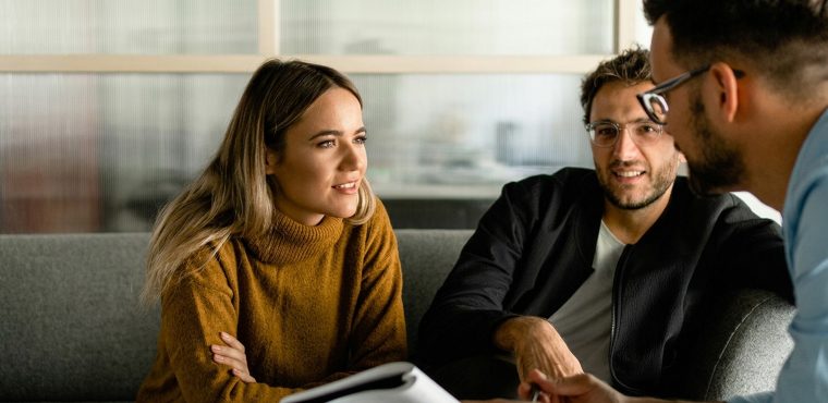 Three professional team members, including a woman in a mustard-yellow turtleneck sweater, sitting on a grey couch collaborating and discussing user-generated content strategies for a community feedback loop