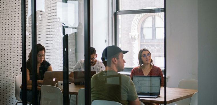 A corporate marketing team working on laptops around a wooden conference table, seen through a glass office partition, illustrating an enterprise transition from single-channel SEO to AI growth engines