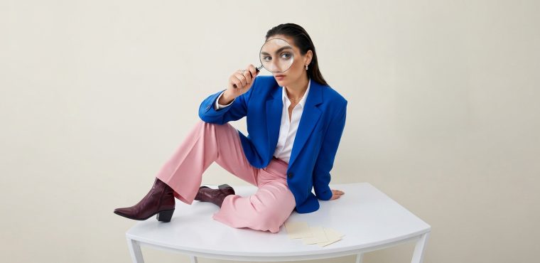 A female marketing analyst in a vibrant royal blue blazer, white collared shirt, light pink pants, and maroon boots sitting atop a white table holding a magnifying glass up to her eye, analyzing the direct relationship between traditional SEO and Generative Engine Optimization