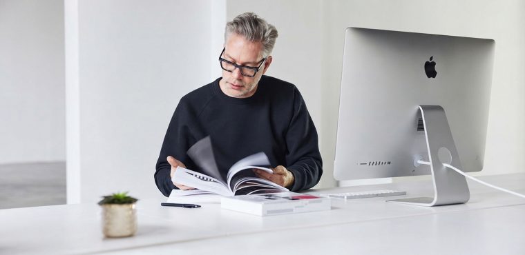 A male content strategist wearing glasses and a black sweatshirt reviewing a printed document next to an iMac desktop computer on a clean white desk, conducting an entity gap analysis for better search rankings