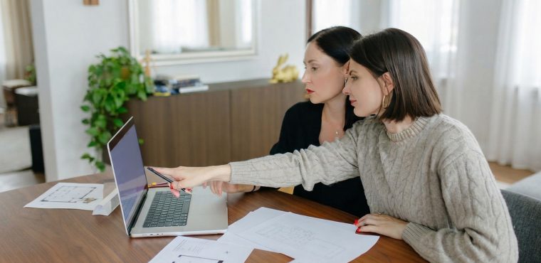 Two female business consultants collaborating at a wooden table over a laptop screen and printed floor plan schematics, illustrating why Gemini Enterprise for CX requires Generative Engine Optimization