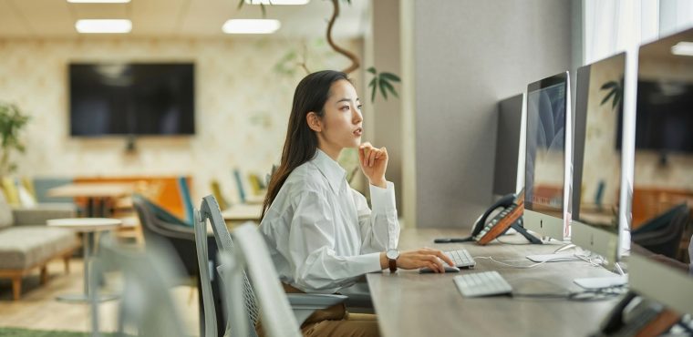 An Asian female SEO strategist in a crisp white shirt contemplating data on desktop monitors in a modern office, planning Generative Engine Optimization (GEO) strategies to replace keyword density with semantic entity density