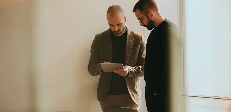 Two male marketing executives—one in a tan corduroy blazer interacting with a digital tablet and one in a black sweater and glasses—reviewing Generative Engine Optimization data for an AI SEO agency in Anaheim, CA