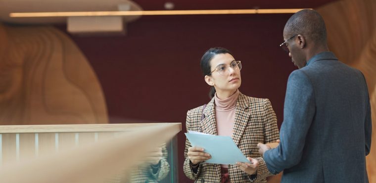 A female corporate executive in a plaid blazer and glasses holding audit reports while discussing Google Tag updates and revenue attribution leaks with a male colleague in a modern office atrium