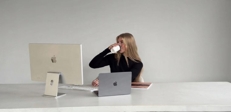 A blonde female corporate strategist in a black top drinking coffee while working at a desk with an Apple iMac and MacBook Pro, illustrating the inverted funnel and buying behavior changes in the agentic AI era