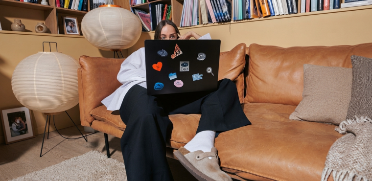 A woman wearing wide-leg trousers, white socks, and suede clogs lounging on a tan leather couch behind a black laptop with colorful stickers, illustrating how AI information synthesis alters the buyer journey