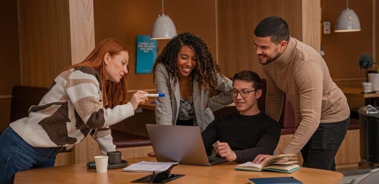 A diverse marketing team of four professionals gathered around a wooden table in a warm modern office, reviewing laptop analytics while a woman holding a whiteboard marker points to key data, illustrating strategic adaptation in the new AI search economy