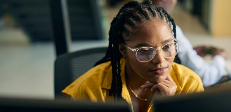 A close-up of a data analyst with braided hair wearing clear-rimmed glasses and a yellow top, leaning forward and focusing deeply on dual computer monitors in a corporate office setting.