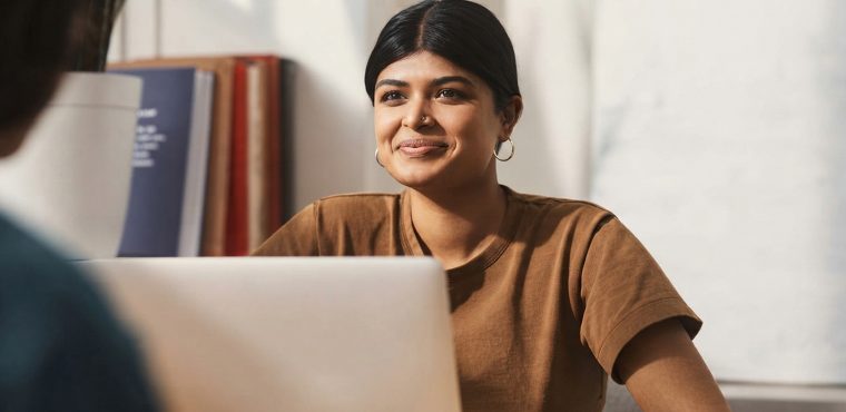 A South Asian female digital marketing strategist in a brown t-shirt smiling behind a laptop while advising a client during a consultation on optimizing for Retrieval-Augmented Generation and AI search citations