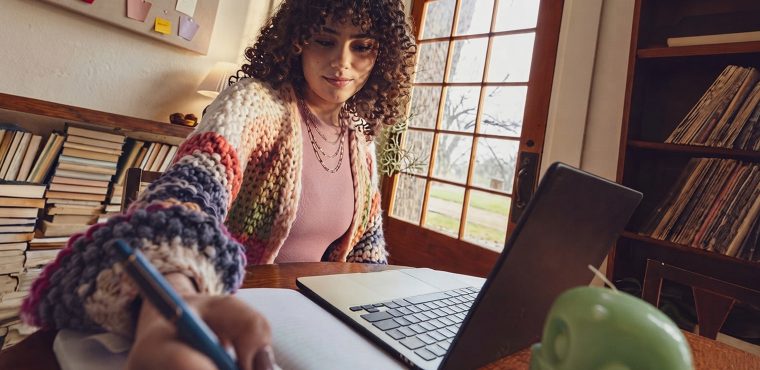 A female enterprise strategy consultant with curly dark hair wearing a colorful knit cardigan writing with a pen in a notebook next to a laptop in a cozy room with bookshelves and French doors, evaluating Orange County GEO agencies for enterprise AI search