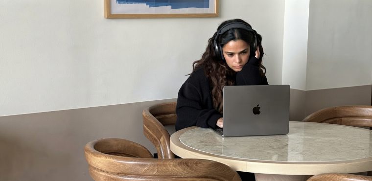 A female digital growth strategist with dark wavy hair wearing black over-ear headphones and a sweater sitting focused at a marble table working on a laptop, comparing traditional SEO and growth infrastructure across Orange County AI marketing agencies