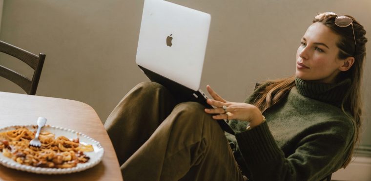 A female digital marketing strategist wearing a green turtleneck sweater and sunglasses pushed up on her head sitting back with her laptop resting on her knees near a dining table, examining Generative Engine Optimization trends replacing traditional SEO for Orange County brands
